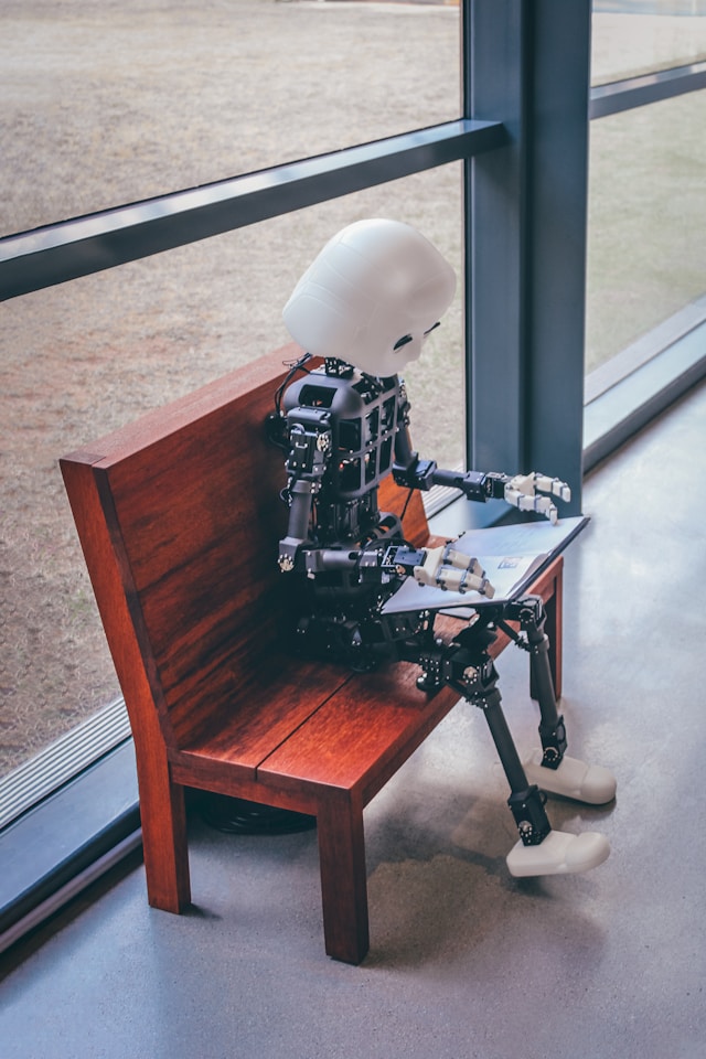 A robot with a white-colored head sits on a bench and looks at a book