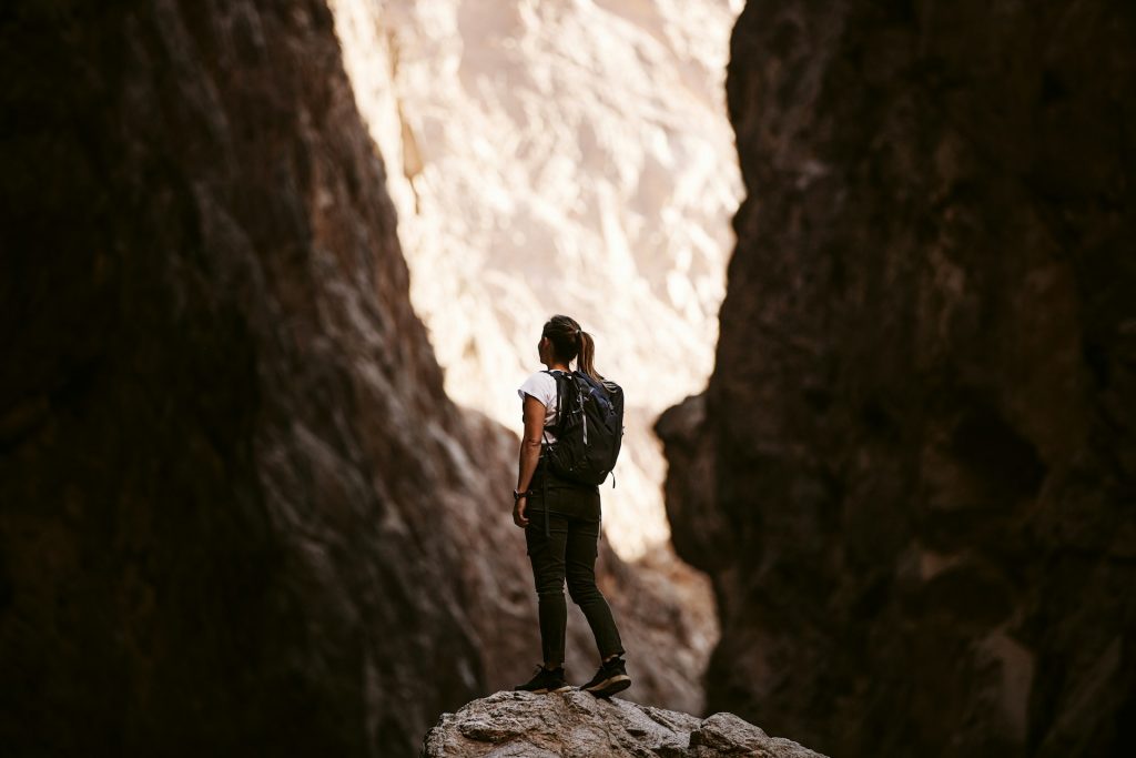 A woman with a white shirt and a black rucksack stands on a rock and looks at a canyon.