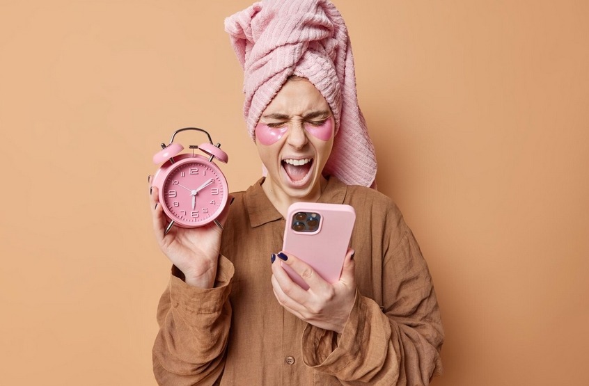 A person with a towel wrapped on their head holds a pink alarm clock and a phone, reacting with surprise or frustration.